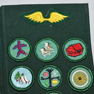 May include: A green fabric sash with a yellow felt winged bird patch and nine round embroidered patches. The patches feature a red starfish, a white and blue ice skate, a red treasure chest, a green tree, a white wagon wheel, and other designs.