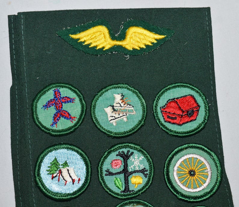 May include: A green fabric sash with a yellow felt winged bird patch and nine round embroidered patches. The patches feature a red starfish, a white and blue ice skate, a red treasure chest, a green tree, a white wagon wheel, and other designs.