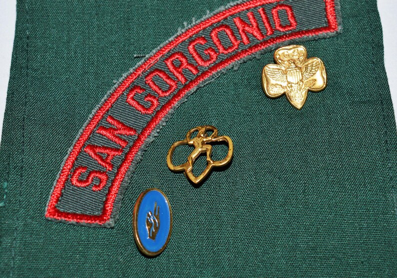 May include: A green fabric patch with red embroidered text that reads "SAN GORGENO". There are three gold-coloured pins attached to the fabric. One pin is a four-leaf clover with a figure in the centre. Another pin is a four-leaf clover with a bird in the centre. The third pin is a blue circle with a white symbol.