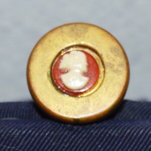 May include: A gold-colored round button with a red and white cameo of a woman's profile.