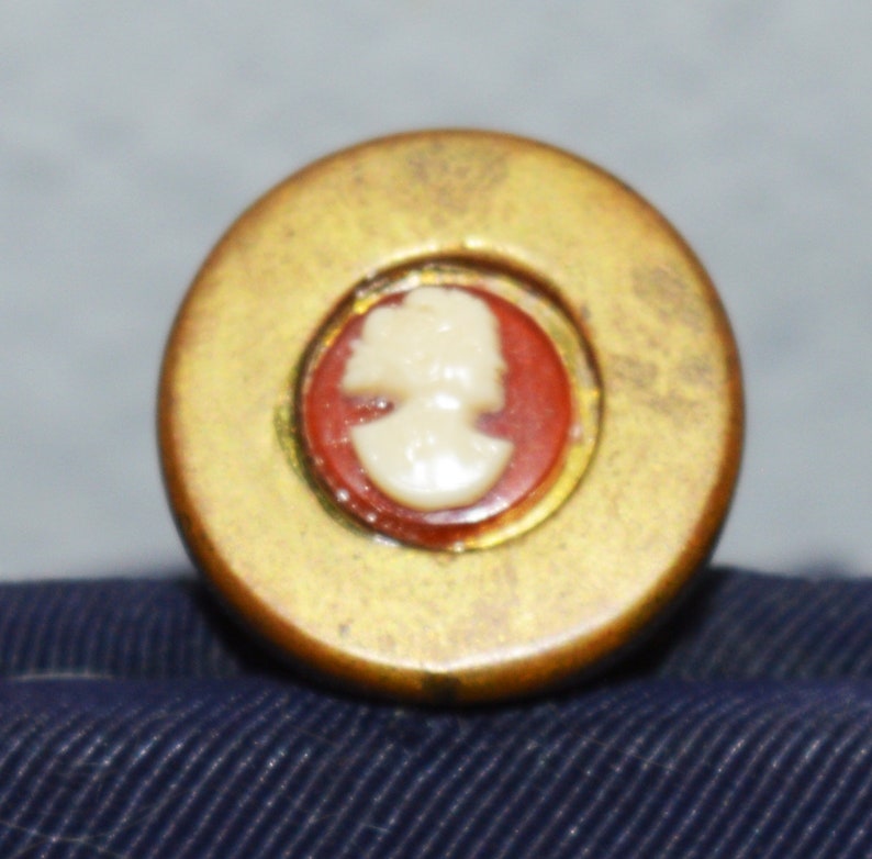 May include: A gold-colored round button with a red and white cameo of a woman's profile.