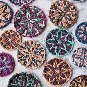 May include: A collection of 14 crocheted mandala coasters in various colours, including pink, green, brown, blue, and grey. The coasters feature intricate geometric patterns and are arranged on a light grey surface.