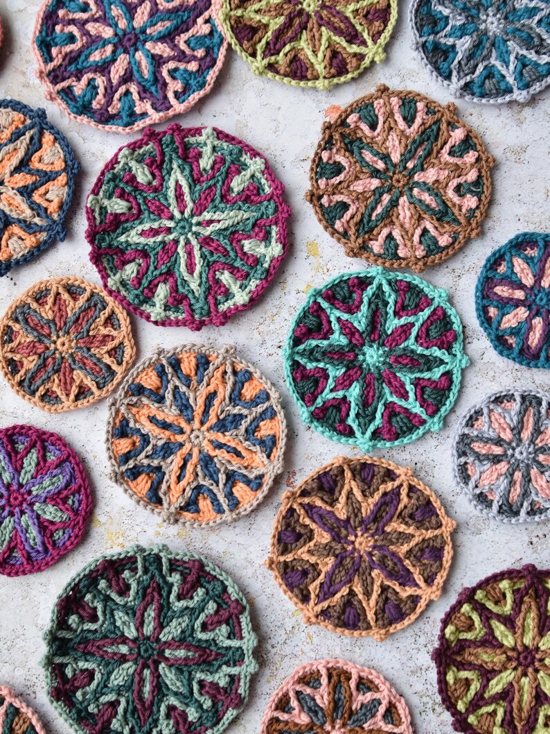 May include: A collection of 14 crocheted mandala coasters in various colours, including pink, green, brown, blue, and grey. The coasters feature intricate geometric patterns and are arranged on a light grey surface.