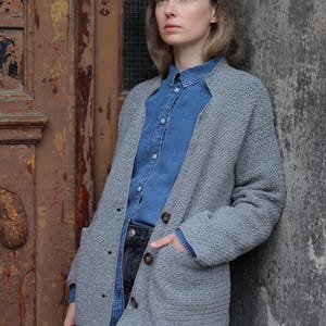 Op de afbeelding: Een grijze gebreide cardigan met knoopsluiting en een zak aan de linkerkant. De cardigan wordt gedragen over een blauw denim shirt en een zwarte jeans.