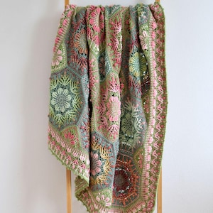 May include: A handmade crochet blanket featuring a complex assembly of hexagonal and floral motifs in shades of sage green, dusty pink, and muted teal. The textile displays intricate yarn work with raised textures and a scalloped border edge.