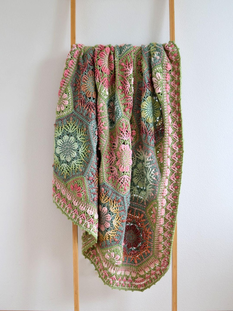 May include: A handmade crochet blanket featuring a complex assembly of hexagonal and floral motifs in shades of sage green, dusty pink, and muted teal. The textile displays intricate yarn work with raised textures and a scalloped border edge.