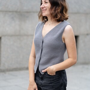 Op de afbeelding: Een grijze, mouwloze gehaakte vest met een V-hals en knoopsluiting. Het vest wordt gedragen met een zwarte jeans. De vrouw glimlacht en kijkt opzij. Het vest is gemaakt van gebreide stof.