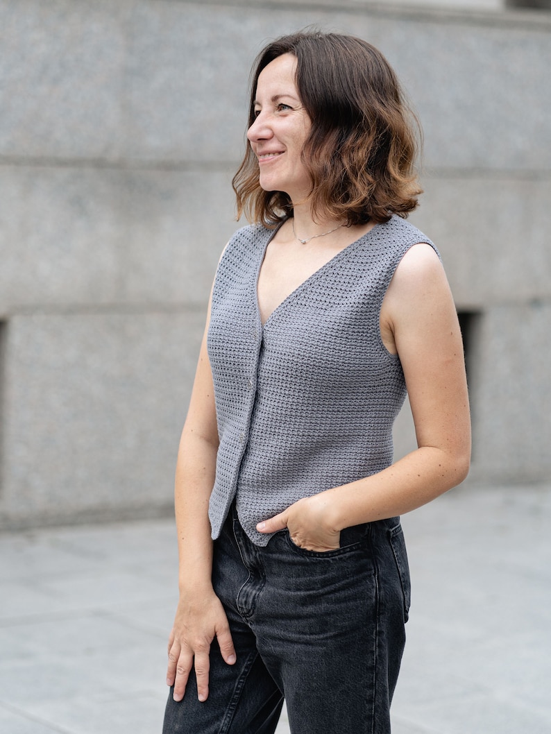 Op de afbeelding: Een grijze, mouwloze gehaakte vest met een V-hals en knoopsluiting. Het vest wordt gedragen met een zwarte jeans. De vrouw glimlacht en kijkt opzij. Het vest is gemaakt van gebreide stof.