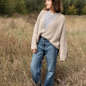 Può includere: Una donna indossa un cardigan beige a trecce sopra una maglia a righe e jeans blu, in piedi in un campo di erba alta. Il cardigan ha un design avvolgente e maniche lunghe. I jeans sono di un lavaggio chiaro.