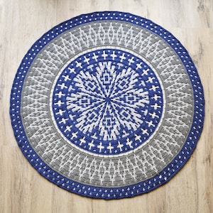 Può includere: Un tappeto rotondo all'uncinetto con un motivo geometrico blu e grigio. Il tappeto ha un design circolare con un medaglione centrale e anelli concentrici di forme geometriche.