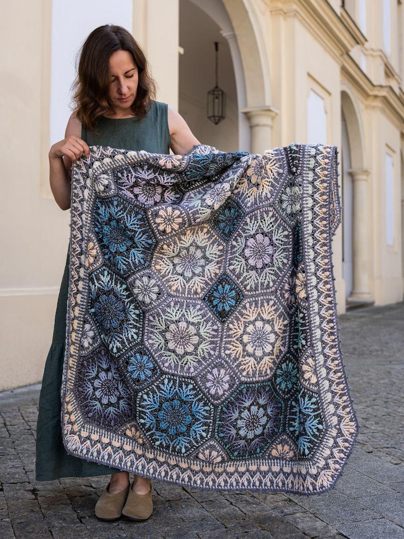 May include: A large, rectangular crochet blanket featuring a complex pattern of interconnected hexagonal motifs. The design incorporates shades of deep blue, slate gray, soft lavender, and cream, with intricate floral-inspired textures.