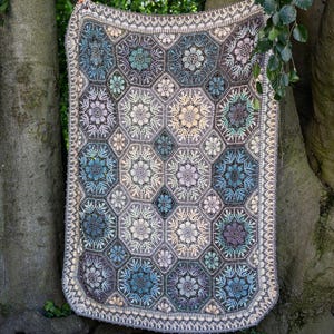 May include: A rectangular crochet blanket featuring a repeating pattern of hexagonal motifs. Each hexagon contains a central floral design in shades of blue, purple, cream, and grey. The blanket is finished with a decorative cream-colored border.