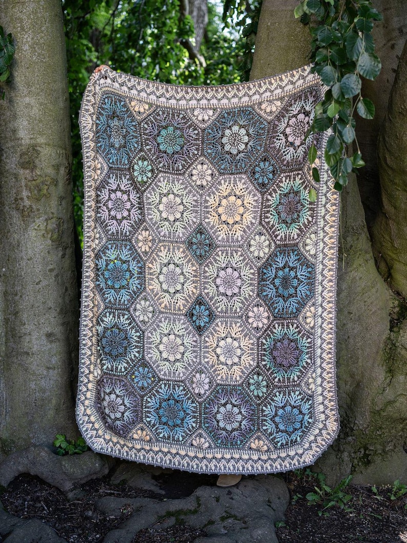 May include: A rectangular crochet blanket featuring a repeating pattern of hexagonal motifs. Each hexagon contains a central floral design in shades of blue, purple, cream, and grey. The blanket is finished with a decorative cream-colored border.