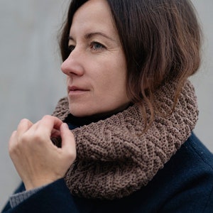 May include: A woman wearing a brown knitted cowl neck scarf and a dark blue coat. She is looking off to the side.