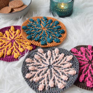 May include: A collection of colorful, handmade crochet coasters. The coasters feature intricate floral designs in various colors, including teal, yellow, pink, and gray. A teal candle holder and a bowl of cookies are also in the image.