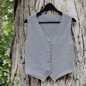 Op de afbeelding: Een grijze, gehaakte vest met een V-hals en drie decoratieve knopen. De vest wordt op een zwarte hanger gepresenteerd tegen een boomstamachtergrond. De vest heeft een neutrale kleur en lijkt handgemaakt.