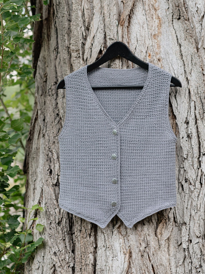 Op de afbeelding: Een grijze, gehaakte vest met een V-hals en drie decoratieve knopen. De vest wordt op een zwarte hanger gepresenteerd tegen een boomstamachtergrond. De vest heeft een neutrale kleur en lijkt handgemaakt.