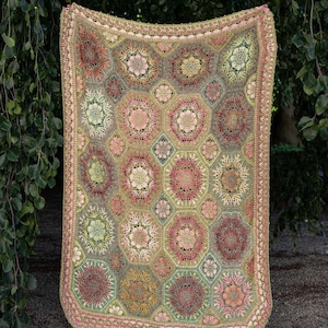 May include: A rectangular crochet blanket featuring a repeating pattern of hexagonal and diamond-shaped motifs in shades of sage green, dusty rose, mustard yellow, and cream. The edges are finished with a decorative scalloped crochet border.