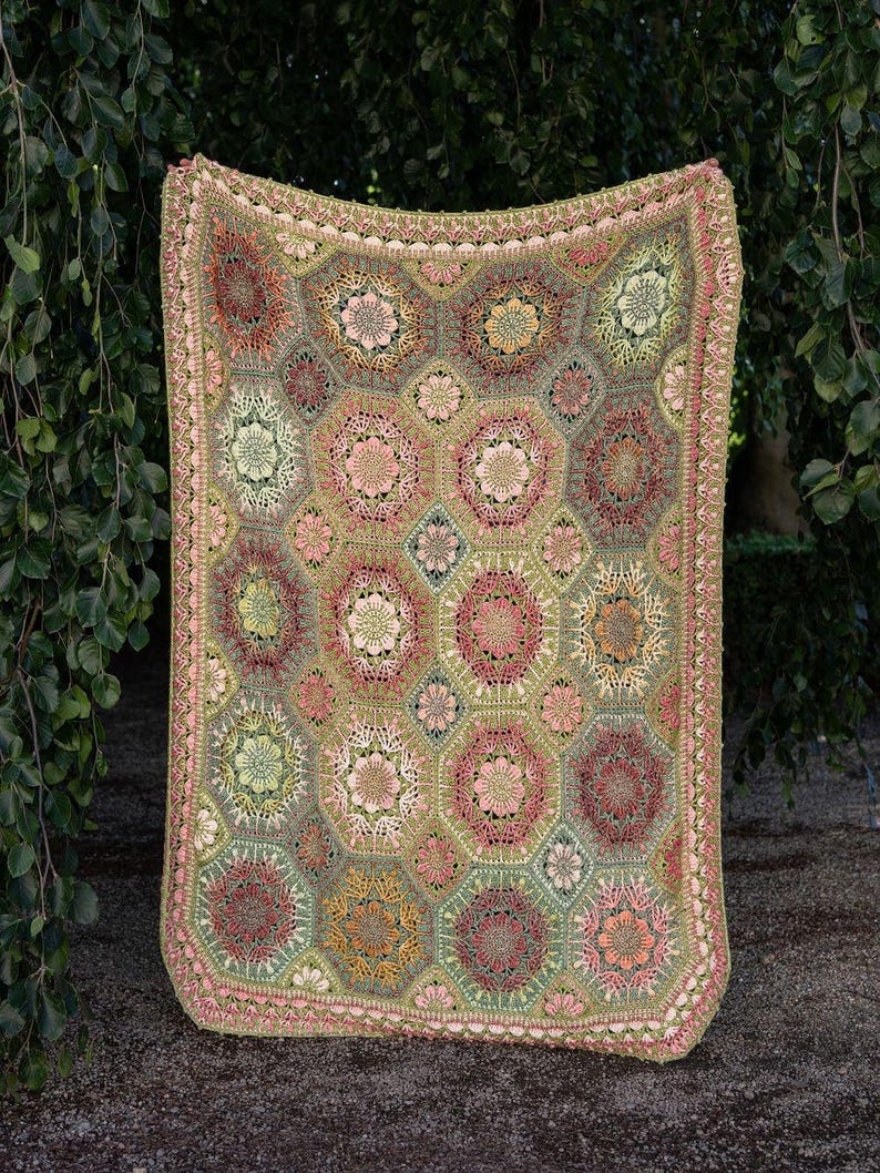 May include: A rectangular crochet blanket featuring a repeating pattern of hexagonal and diamond-shaped motifs in shades of sage green, dusty rose, mustard yellow, and cream. The edges are finished with a decorative scalloped crochet border.