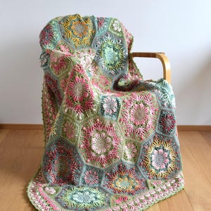 May include: A handmade crochet blanket composed of interconnected hexagonal motifs featuring intricate floral patterns in shades of sage green, dusty pink, mustard yellow, and cream. The textured design displays raised stitches and botanical shapes.