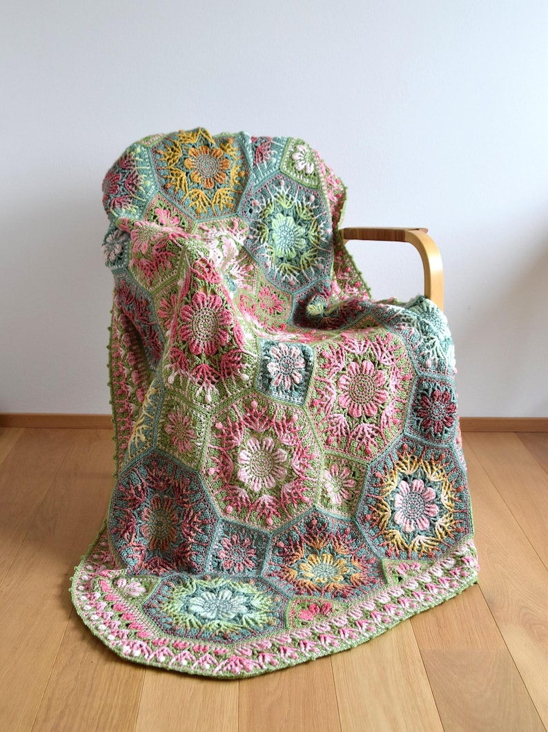 May include: A handmade crochet blanket composed of interconnected hexagonal motifs featuring intricate floral patterns in shades of sage green, dusty pink, mustard yellow, and cream. The textured design displays raised stitches and botanical shapes.