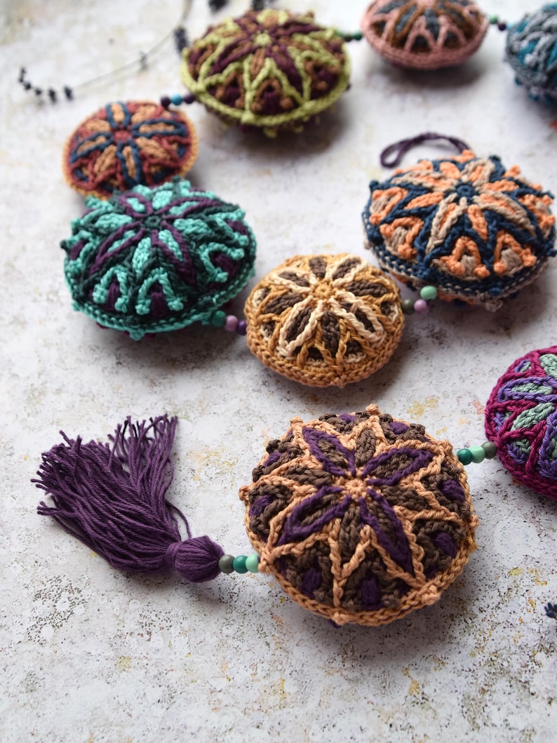 May include: A garland of crocheted round ornaments with intricate geometric patterns in shades of brown, green, blue, and purple. The ornaments are strung together with beads and a purple tassel.