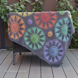 Könnte beinhalten: Eine bunte Häkeldecke mit einem geometrischen Muster aus Achtecken und Kreisen. Die Decke ist in verschiedenen Farben gehalten, darunter Orange, Gelb, Grün, Blau und Lila. Die Decke liegt über einem schwarzen Stuhl.