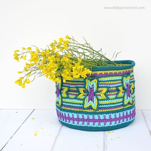 Può includere: Un cesto di crochet color turchese, viola e giallo con un motivo geometrico. Il cesto è pieno di fiori gialli.