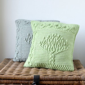 Puede incluir: Dos fundas de cojín de crochet con un diseño de árbol. Una es verde y la otra es gris. Los cojines están sobre una superficie de mimbre.