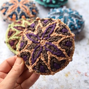 May include: A set of crocheted coasters in various colours and patterns. The coasters are round and feature a geometric design. The coasters are made with brown, purple, green, blue, and orange yarn.