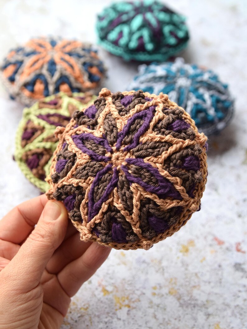 May include: A set of crocheted coasters in various colours and patterns. The coasters are round and feature a geometric design. The coasters are made with brown, purple, green, blue, and orange yarn.