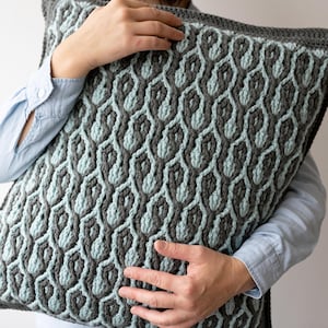 May include: A crocheted pillow with a geometric pattern in shades of gray and blue. The pillow is being held by a person.