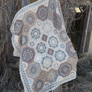 May include: A crochet blanket featuring a repeating pattern of large hexagonal motifs and smaller square connectors. The design incorporates intricate lace-like stitching in a color palette of muted blue, soft brown, cream, and off-white tones.