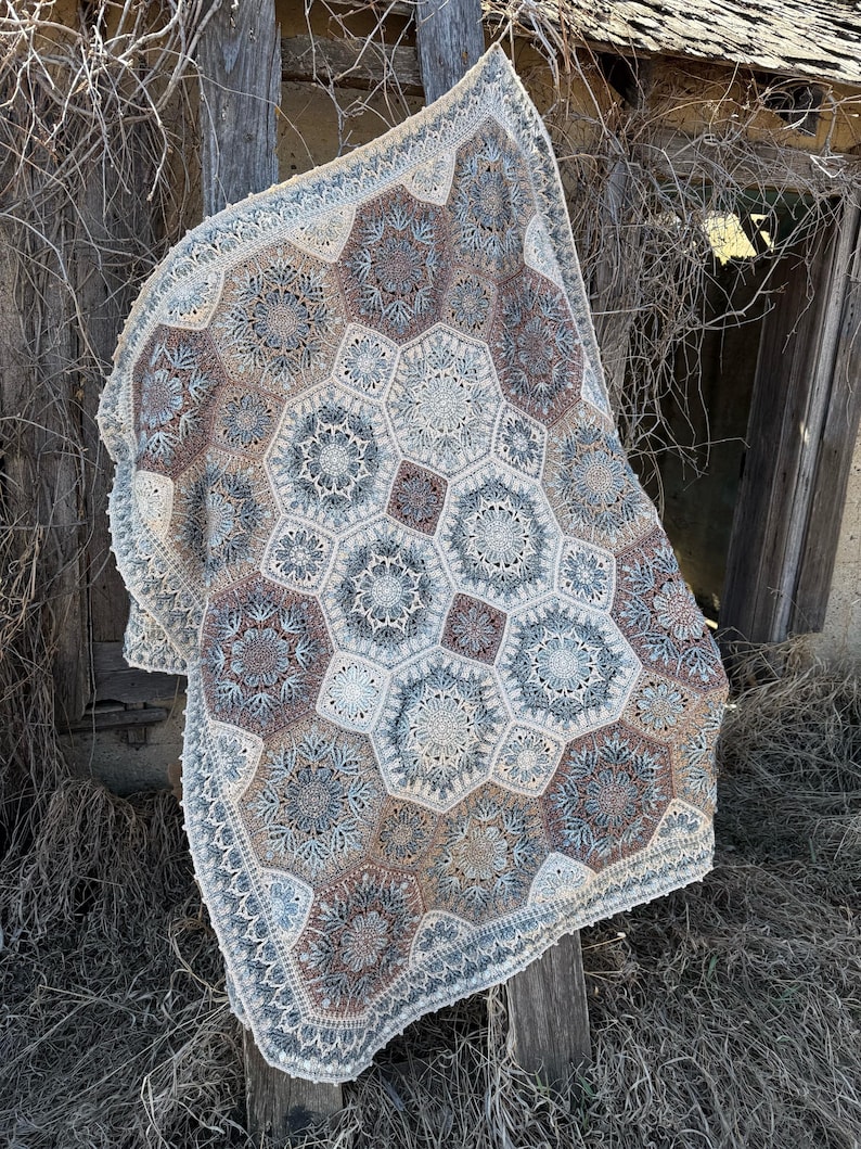 May include: A crochet blanket featuring a repeating pattern of large hexagonal motifs and smaller square connectors. The design incorporates intricate lace-like stitching in a color palette of muted blue, soft brown, cream, and off-white tones.