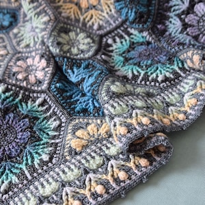 May include: A crochet blanket featuring a series of interconnected hexagonal motifs. The design incorporates floral patterns in shades of dark gray, teal, light blue, lavender, and pale yellow, finished with a decorative border of small bobbles.