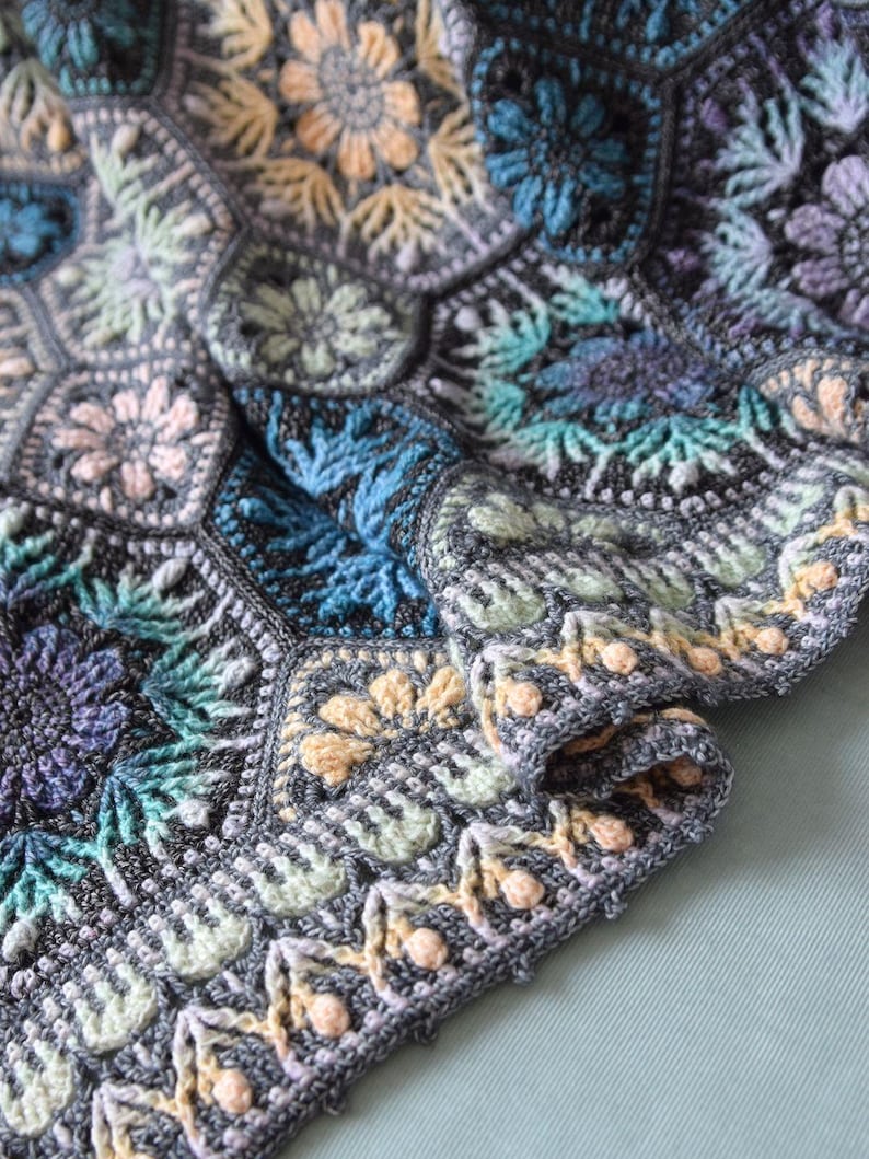 May include: A crochet blanket featuring a series of interconnected hexagonal motifs. The design incorporates floral patterns in shades of dark gray, teal, light blue, lavender, and pale yellow, finished with a decorative border of small bobbles.