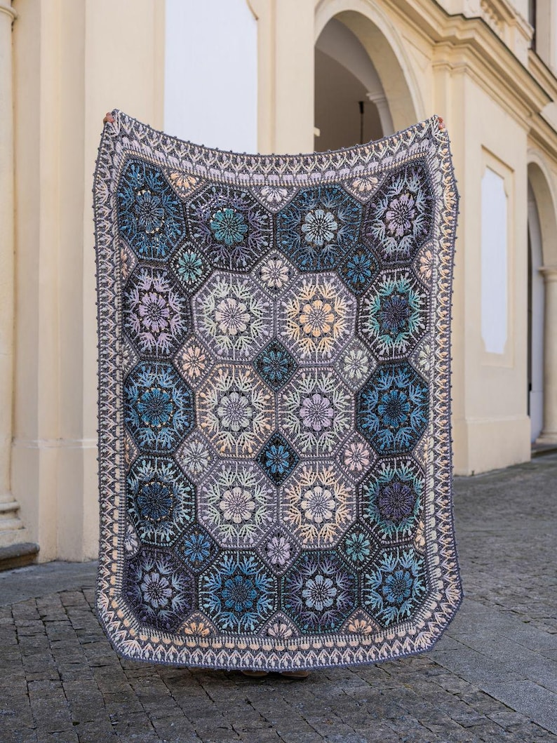 May include: A rectangular crochet blanket featuring a repeating pattern of hexagonal motifs. The design incorporates shades of blue, purple, grey, and cream, with intricate floral-like textures within each hexagon and a decorative border.
