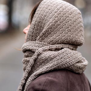 Pode incluir: Uma echarpe com capuz de cor castanho feita de croch&ecirc; com um padr&atilde;o texturizado. A echarpe &eacute; usada sobre a cabe&ccedil;a e o pesco&ccedil;o de uma pessoa, cobrindo o rosto.