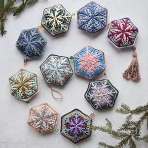 Op de afbeelding: Een slinger van gehaakte sneeuwvlokversieringen in verschillende kleuren, waaronder blauw, groen, roze en bruin. De versieringen zijn met kralen verbonden en aan een van de versieringen hangt een kwast.