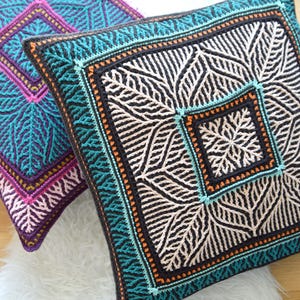 Op de afbeelding: Twee vierkante gehaakte kussens met ingewikkelde bladerpatronen. Eén kussen heeft teal, zwarte en crèmekleuren, met oranje en turquoise accenten. Het andere kussen heeft paarse, roze en turquoise kleuren. Beide liggen op een witte, pluizige ondergrond.