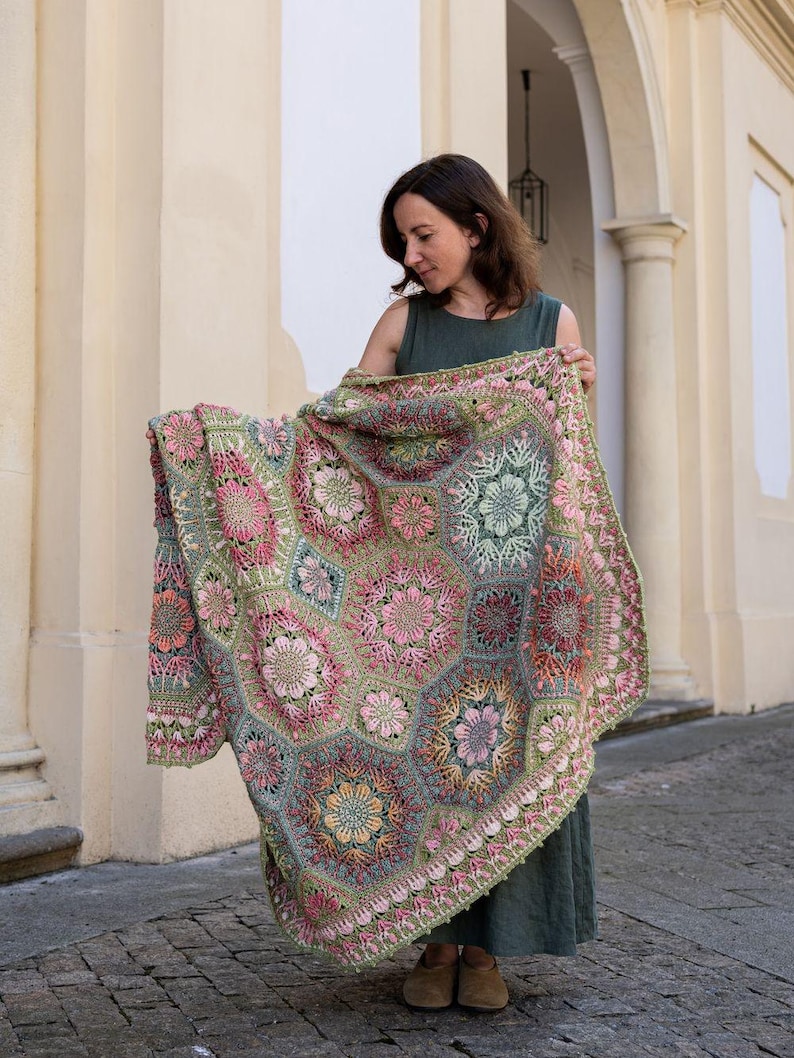 May include: A large, rectangular crochet blanket featuring a complex assembly of hexagonal and floral motifs in shades of sage green, dusty pink, coral, and cream. The intricate needlework displays varied textures and geometric patterns throughout.