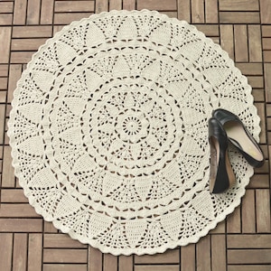 May include: A large, round, crocheted rug in a cream color with a delicate, intricate pattern. The rug is made of yarn and has a circular design with multiple layers of crocheted lace.