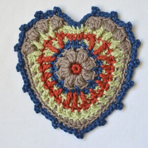 Puede incluir: Un posavasos de coraz&oacute;n tejido a crochet con un borde azul, un centro marr&oacute;n, rojo y verde, y un dise&ntilde;o de flor.