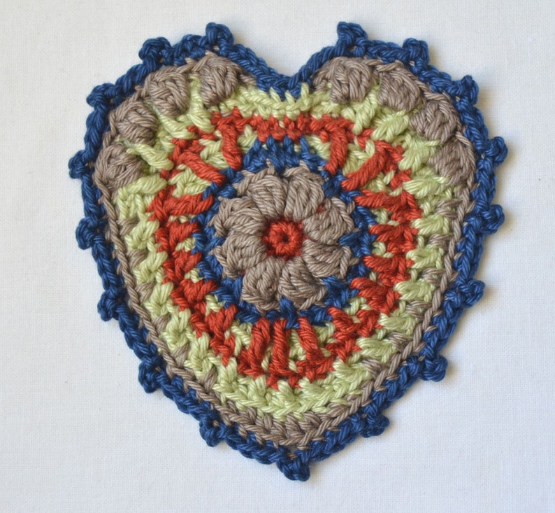 Puede incluir: Un posavasos de coraz&oacute;n tejido a crochet con un borde azul, un centro marr&oacute;n, rojo y verde, y un dise&ntilde;o de flor.