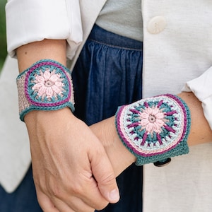 May include: Two handmade crochet wrist cuffs. Each cuff features a central floral design in pink, teal, and purple, surrounded by a teal border. The cuffs are worn on the wrists, showcasing the intricate crochet work and color palette.