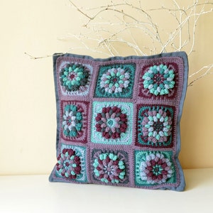 May include: A square crocheted cushion with a grey border and a patchwork design of purple, green and blue flowers.