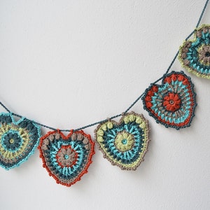 Puede incluir: Una guirnalda de corazones de ganchillo en tonos turquesa, verde, naranja y marr&oacute;n. Los corazones est&aacute;n unidos con un hilo azul oscuro.