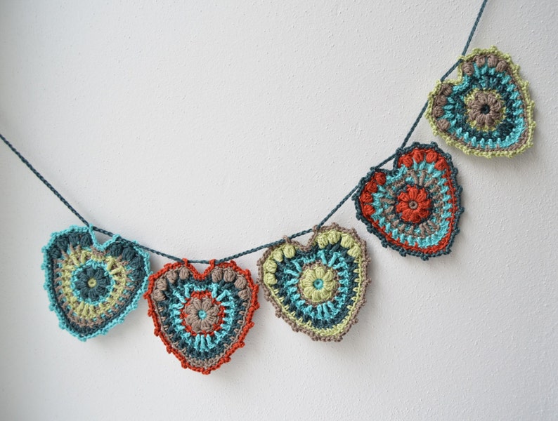 Puede incluir: Una guirnalda de corazones de ganchillo en tonos turquesa, verde, naranja y marr&oacute;n. Los corazones est&aacute;n unidos con un hilo azul oscuro.