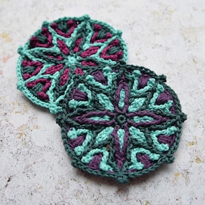 May include: Two crocheted coasters with a geometric pattern in shades of teal and purple.