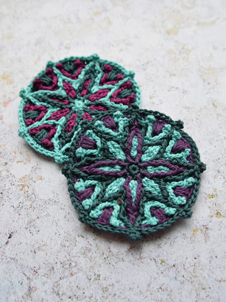 May include: Two crocheted coasters with a geometric pattern in shades of teal and purple.